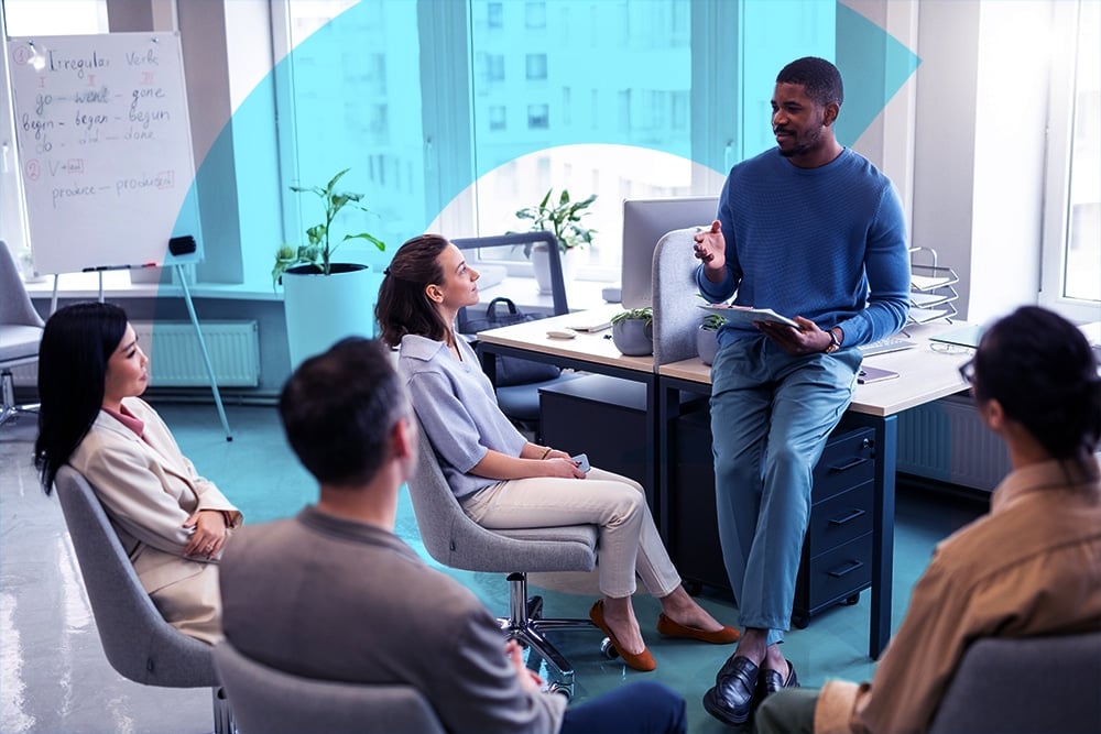 comunicação assertiva - imagem de um homem falando para seus colegas de trabalho, os quais estão sentados ao redor. Sala de escritório com paredes de vidro.